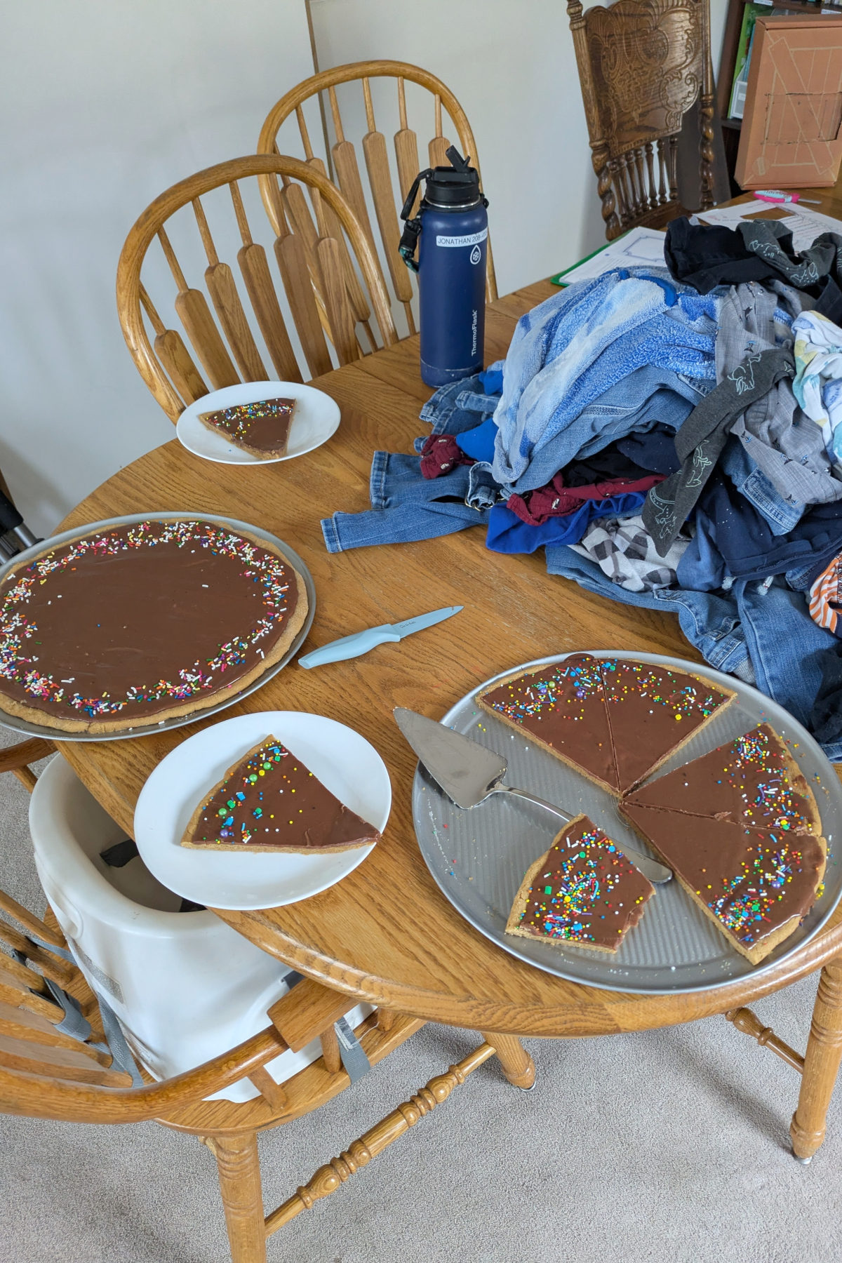 peanut butter cookie cake recipe with chocolate frosting. Two cookie cakes on a table. You can see some cut slices, a knife, and sprinkles on the cakes. There is a pile of laundry next to the cakes.