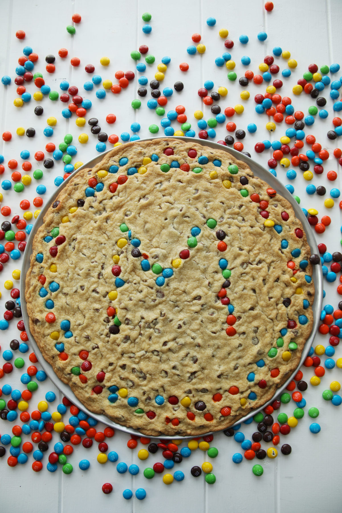 A chocolate chip cookie cake baked in a pizza pan with m and ms on top making an M and a colorful boarder. M and Ms are all over the white table around the cookie.