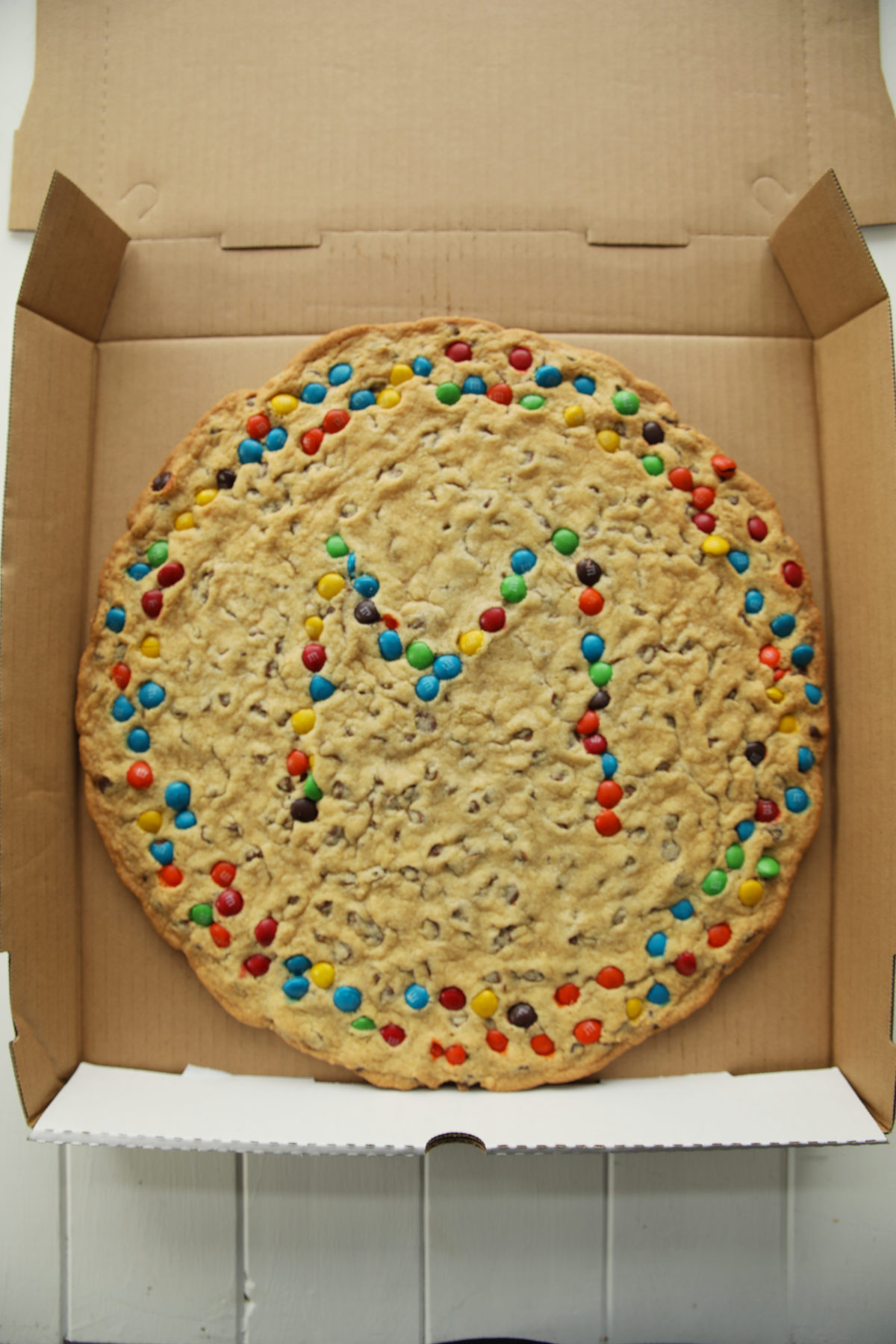 A chocolate chip cookie cake decorated with M and Ms and put into a pizza box ready for delivery.