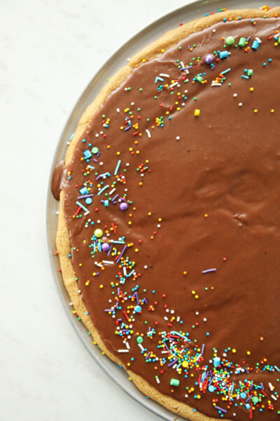 A beautiful peanut butter cookie cake recipe is shown in this image. You can see half the cake in the frame, a light tan with Texas sheet cake frosting and sprinkles around the border.