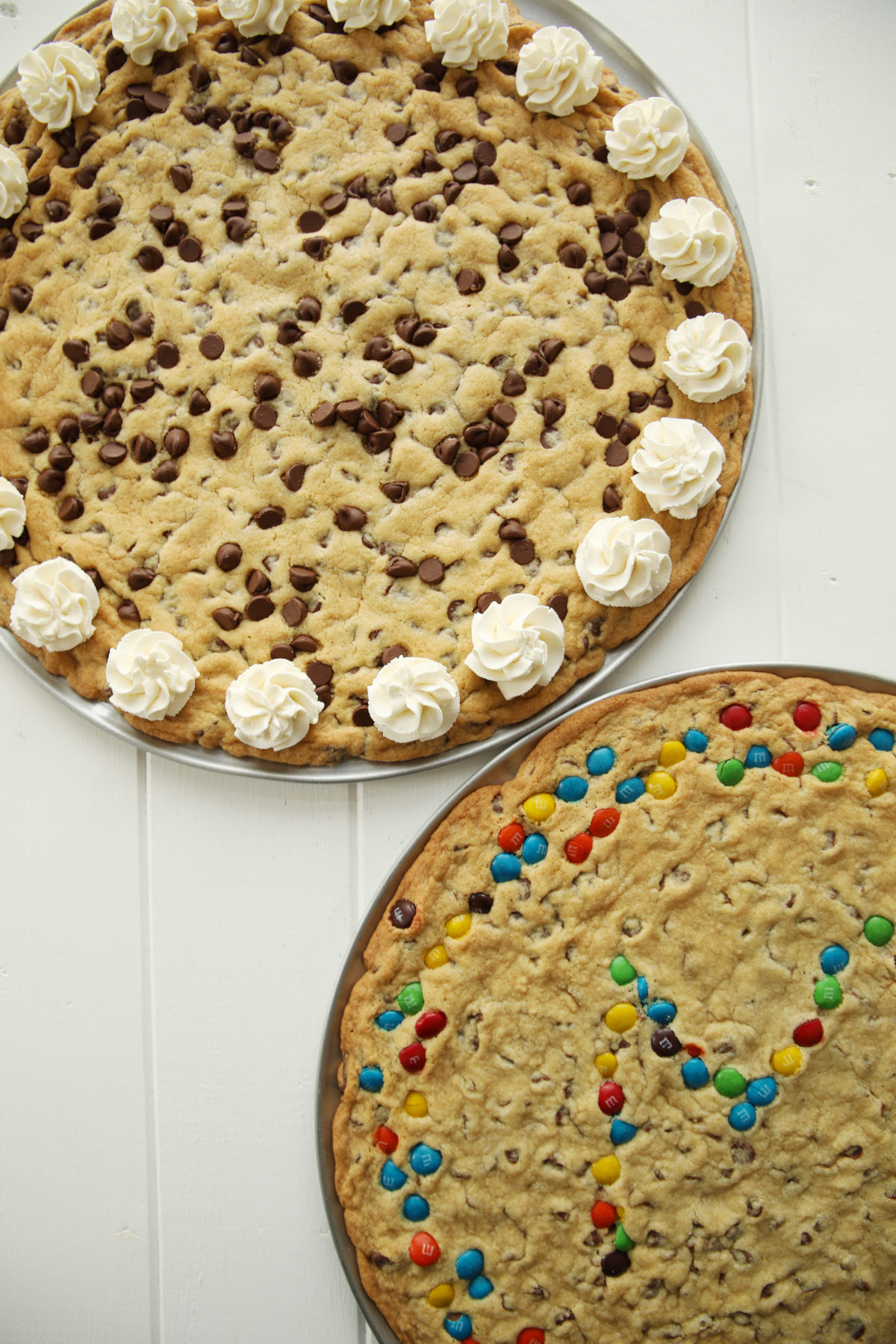 Two chocolate chip cookie cakes on a table. One is decorated with a whipped cream boarder and one is decorated with M&Ms.