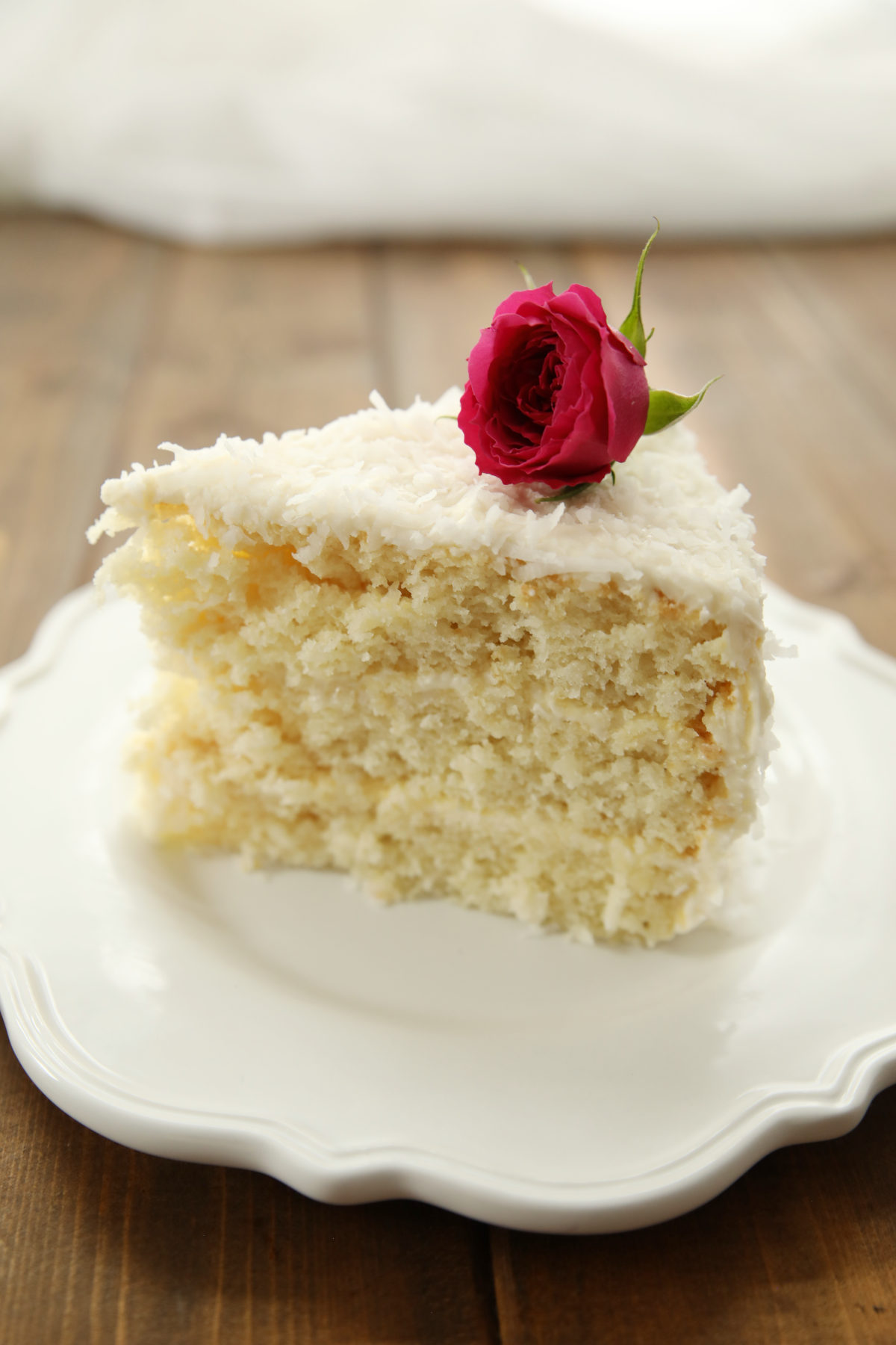 A wedge of coconut cake sits on a white plate. The cake is three layers and has cream cheese frosting and a red rose on top of the coconut cake slice.