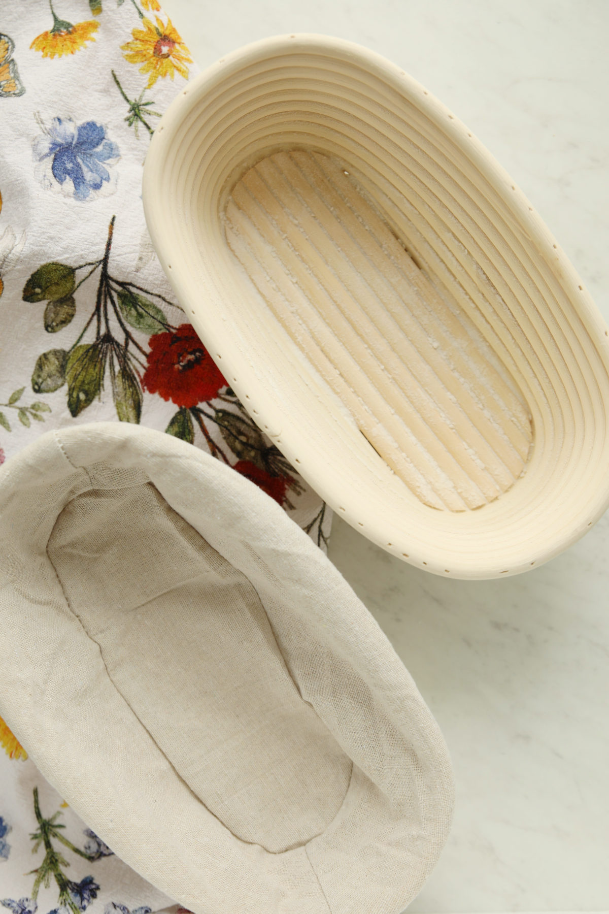 A batard-shaped cane banneton proofing basket and a linen liner on a floral tea towel, an essential tool for sourdough for beginners to help shape bread during the final rise.