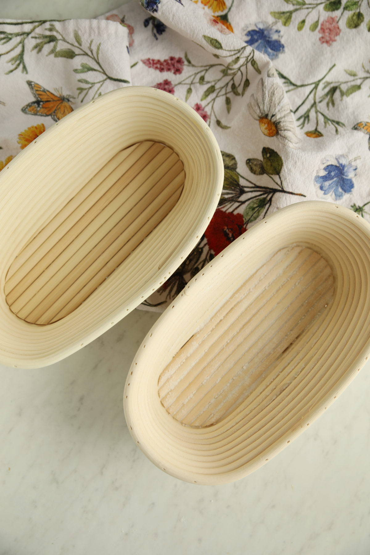 Two oval cane banneton proofing baskets for sourdough baking, sitting on a marble countertop next to a floral tea towel.