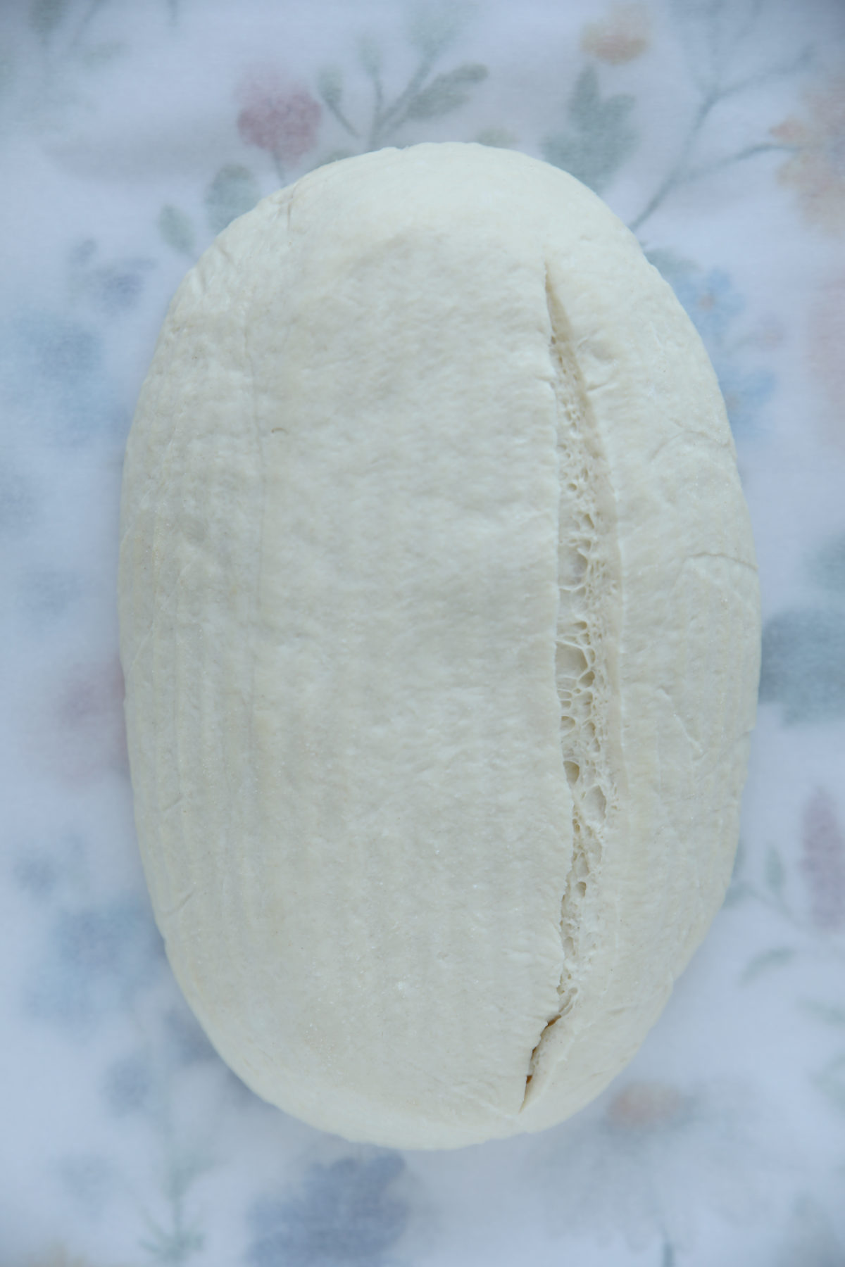 A top-down view of an unbaked sourdough batard on a floral cloth, featuring one long, clean vertical score down the right side of the dough.