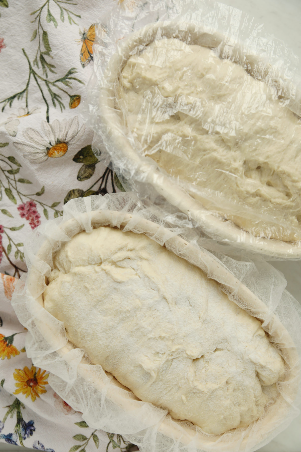Top-down view comparing two oval banneton baskets: one lined with a disposable hairnet showing a rice flour-dusted loaf, and the other covered in plastic wrap.