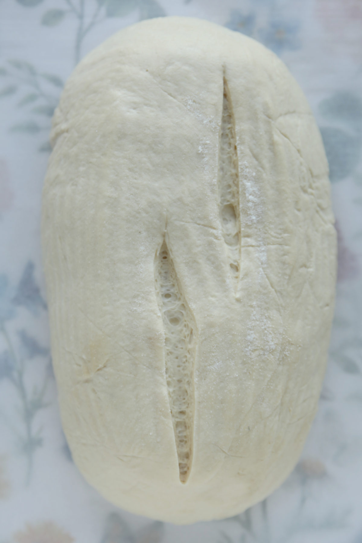 A top-down view of raw sourdough batard dough featuring two vertical, parallel scores down the center, resting on a white floral-patterned tea towel.