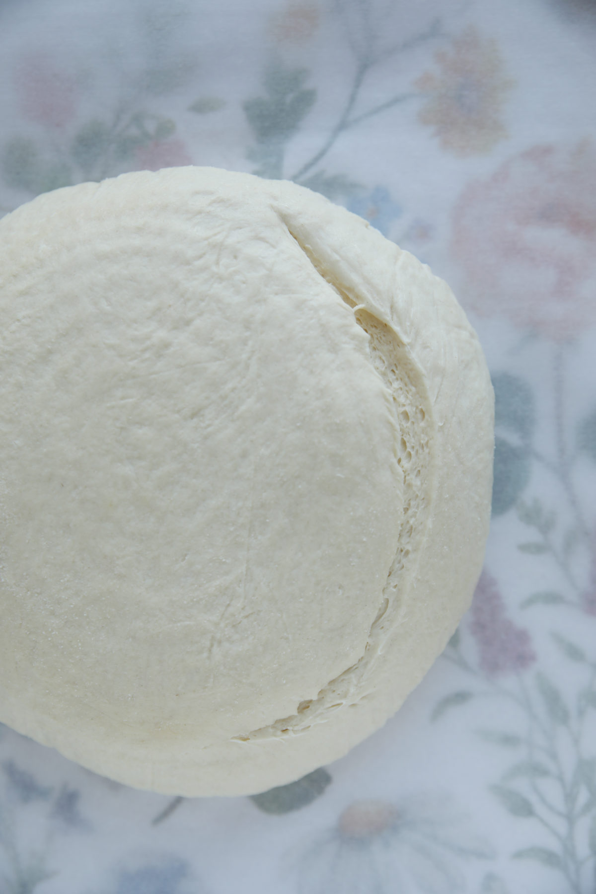A top-down view of a round sourdough boule on a floral tea towel, featuring a single curved, half-moon score along one side of the raw dough.