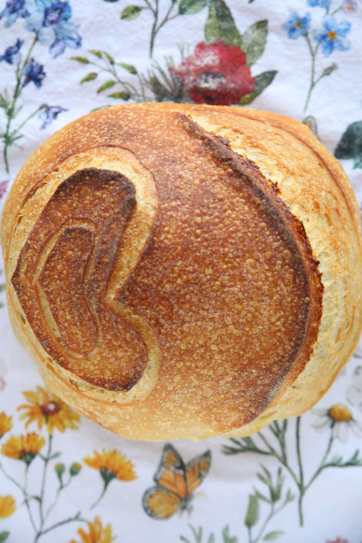 A top-down view of a fully baked, round sourdough boule resting on a floral-patterned cloth. A large heart-shaped score in the center has expanded and browned deeply, creating a rustic, indented outline against the golden-brown crust.
