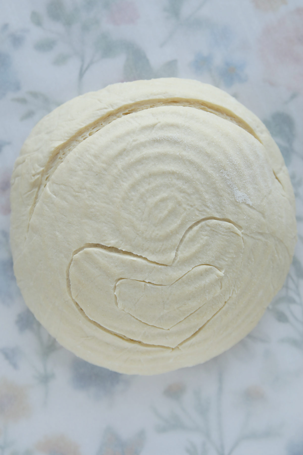 A top-down view of a raw, round sourdough boule featuring a large heart-shaped score outlined in the center of the dough.