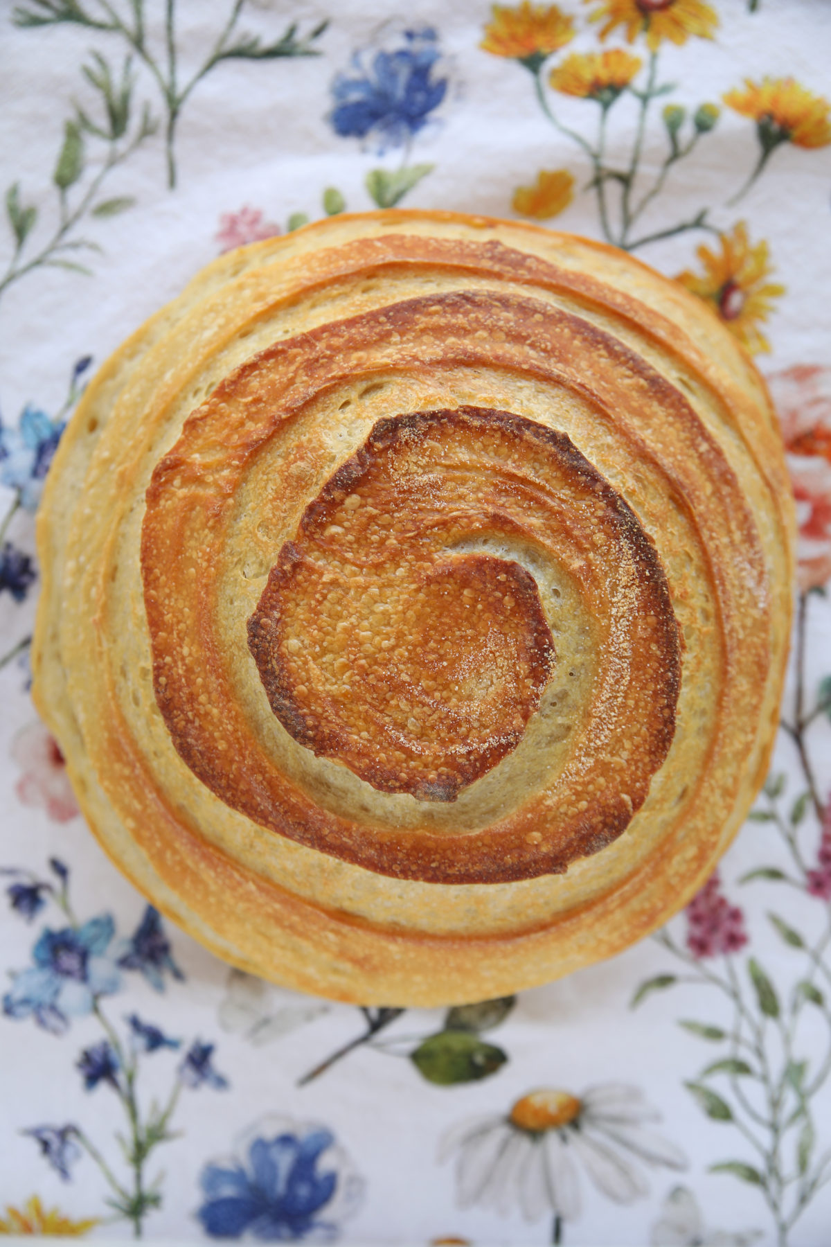 A top-down view of a fully baked, round sourdough boule. The crust is a deep golden brown with a prominent, continuous spiral score that winds from the center out to the edges. The score has opened significantly, showing a textured, toasted interior along the ridges.