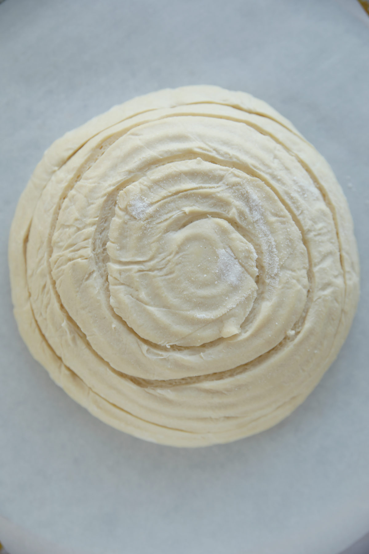 A top-down view of a raw, round sourdough boule featuring a continuous spiral score starting from the center and winding outward to the edges.