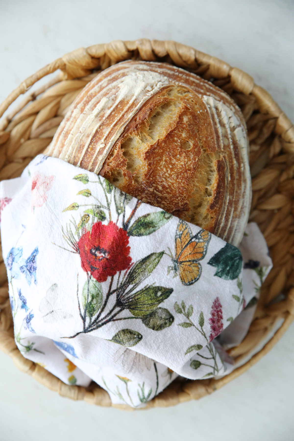 A whole artisanal batard loaf with a prominent ear and flour dusting, tucked into a basket with a floral towel. This is a great example of a rustic finish for sourdough for beginners.