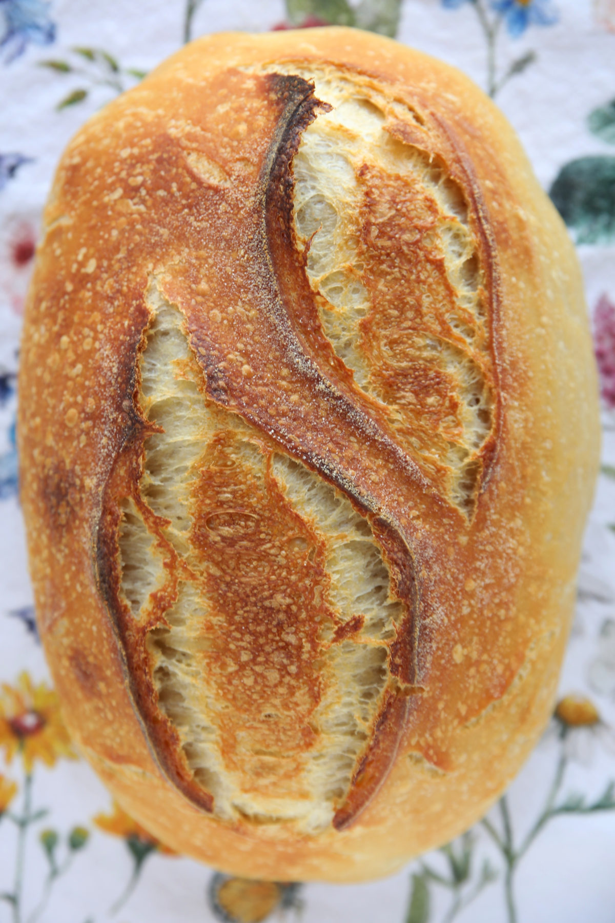 A baked oval sourdough batard with a deep golden-brown crust. Two parallel vertical scores have expanded during baking, creating a wide, symmetrical opening in the center that reveals the lighter-colored, airy bread inside.