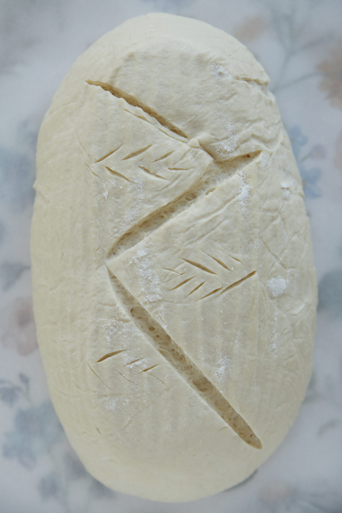 top-down view of a raw sourdough batard on a floral tea towel, scored with a large zig-zag pattern across the center and small decorative wheat-ear slashes inside the triangles.