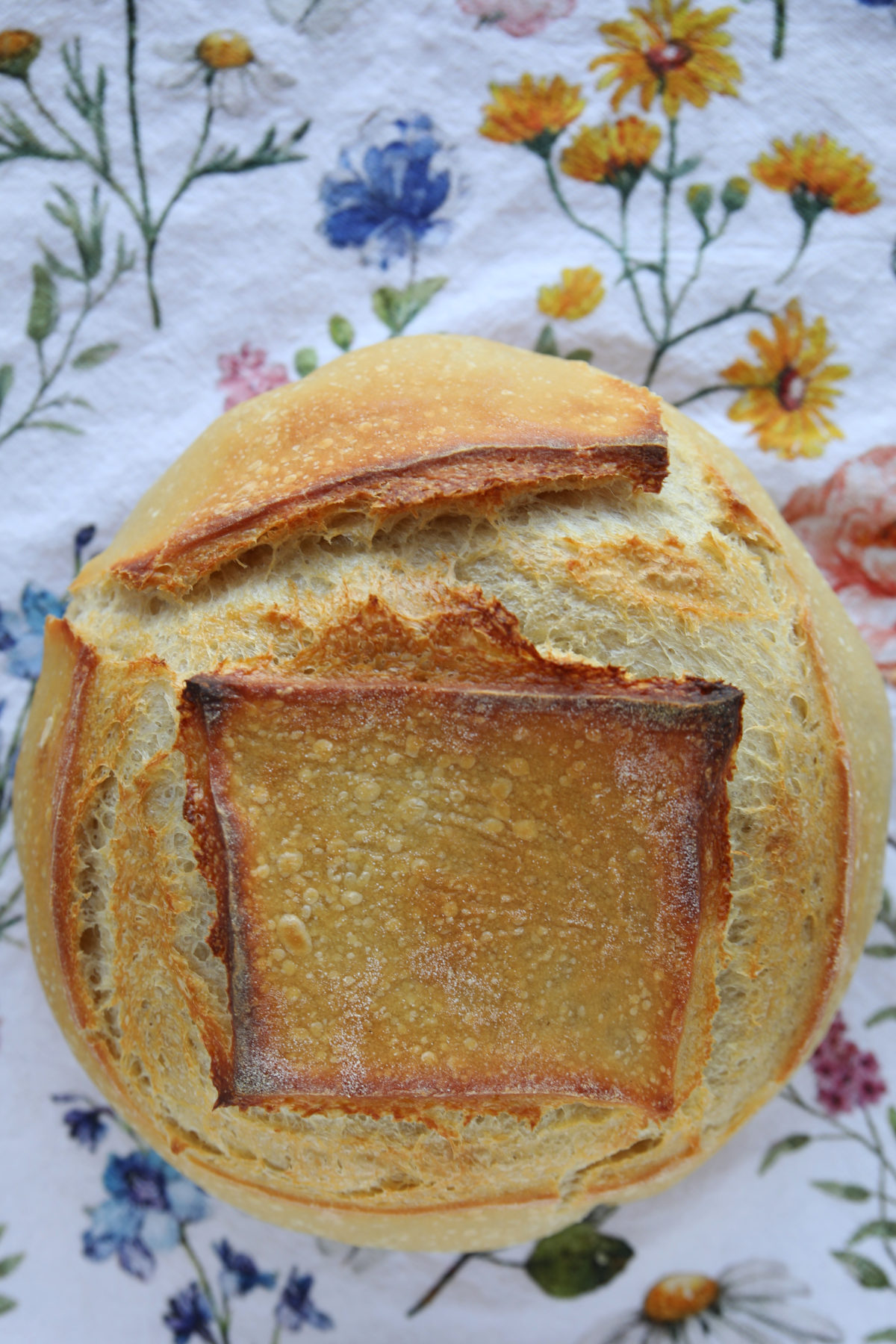 A top-down shot of a golden sourdough loaf with a square scoring pattern that has opened up to reveal a crispy, textured crust, set against a floral background.