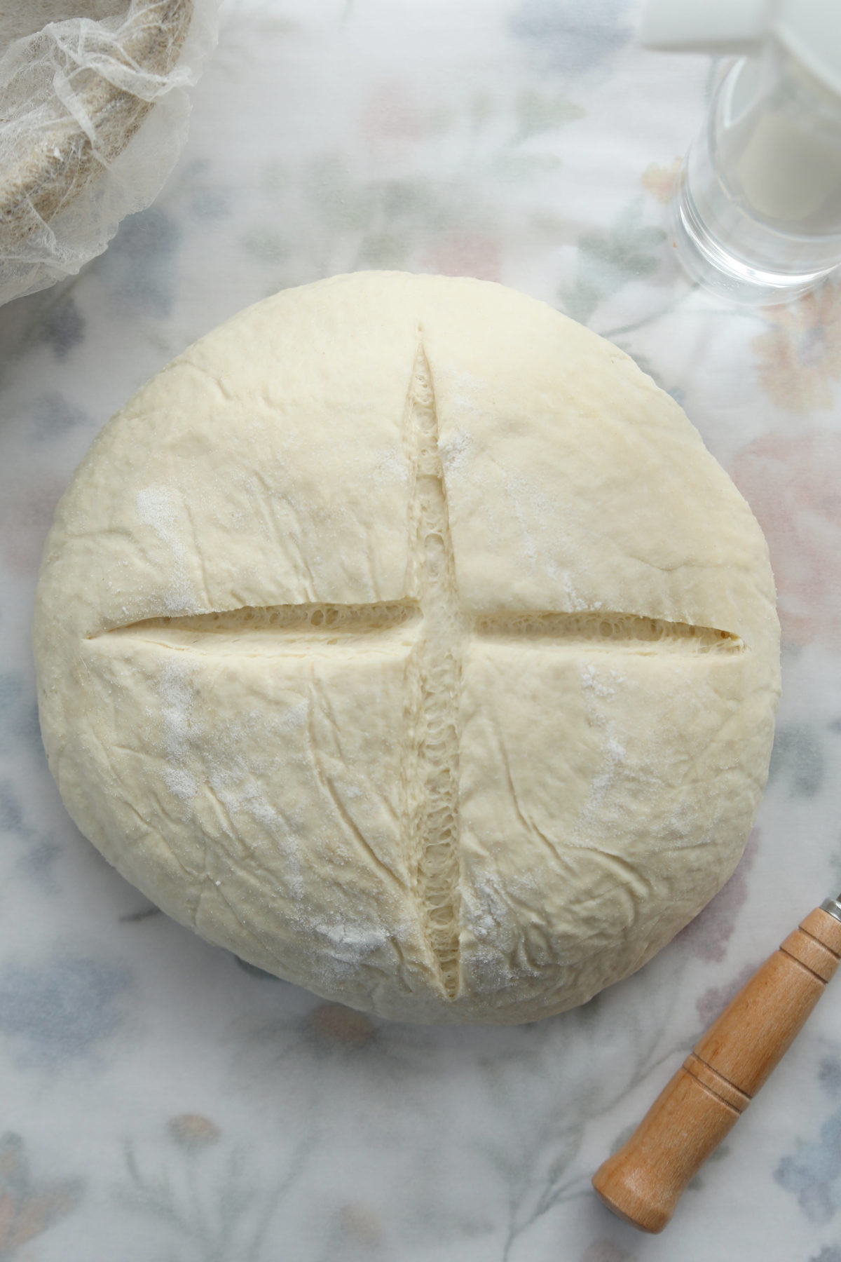 A top-down view of a raw sourdough boule with a deep, symmetrical cross score, ready for the oven.