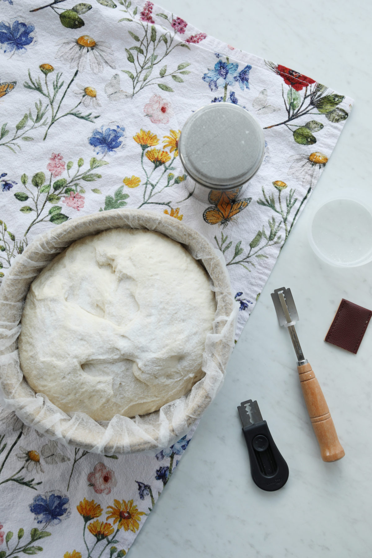 A top-down view of sourdough bread dough proofing in a hair net lined banneton basket on a floral tea towel, surrounded by essential scoring tools including a wood-handled lame, a UFO-style lame, and a flour duster