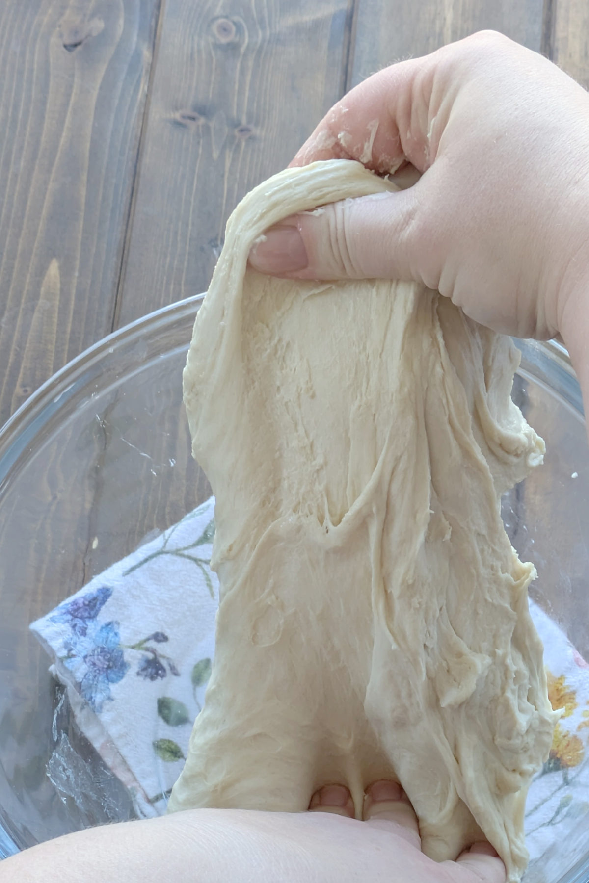 Close-up of hands gently pulling and stretching a pale, elastic ball of sourdough dough upward in a clear glass bowl to perform a stretch and fold.