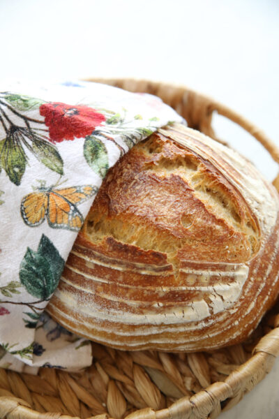 A pretty artisan loaf of sourdough bread is wrapped in a wildflower print tea towel sitting in a basket. This is a decorative image for an article about what sourdough bread is.