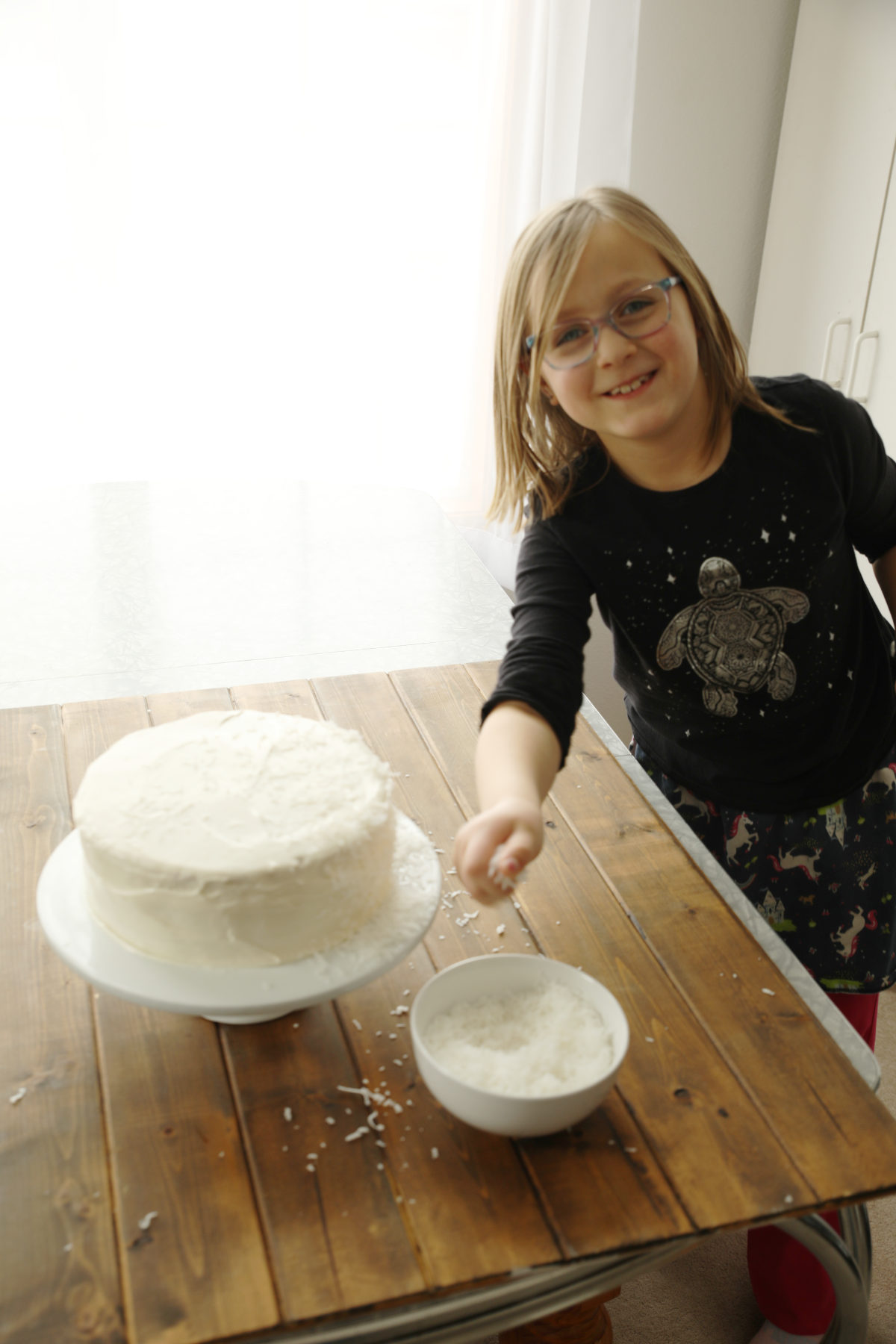 Young girl with glasses throwing shredded coconut at a frosted three-layer coconut cake on a wooden table.