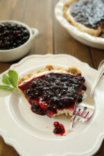 A slice of huckleberry cheesecake is plated on a white plate with a fork and a sprig of mint. You can see the rest of the cheesecake and a white dish of huckleberries in the background.