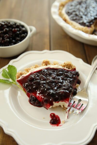 A slice of huckleberry cheesecake is plated on a white plate with a fork and a sprig of mint. You can see the rest of the cheesecake and a white dish of huckleberries in the background.