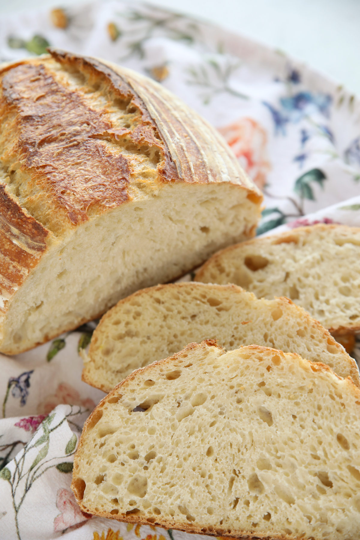 a beautiful loaf of beginning sourdough bread.  The batard shaped bread has three slices in front of it on a floral tea towel. 