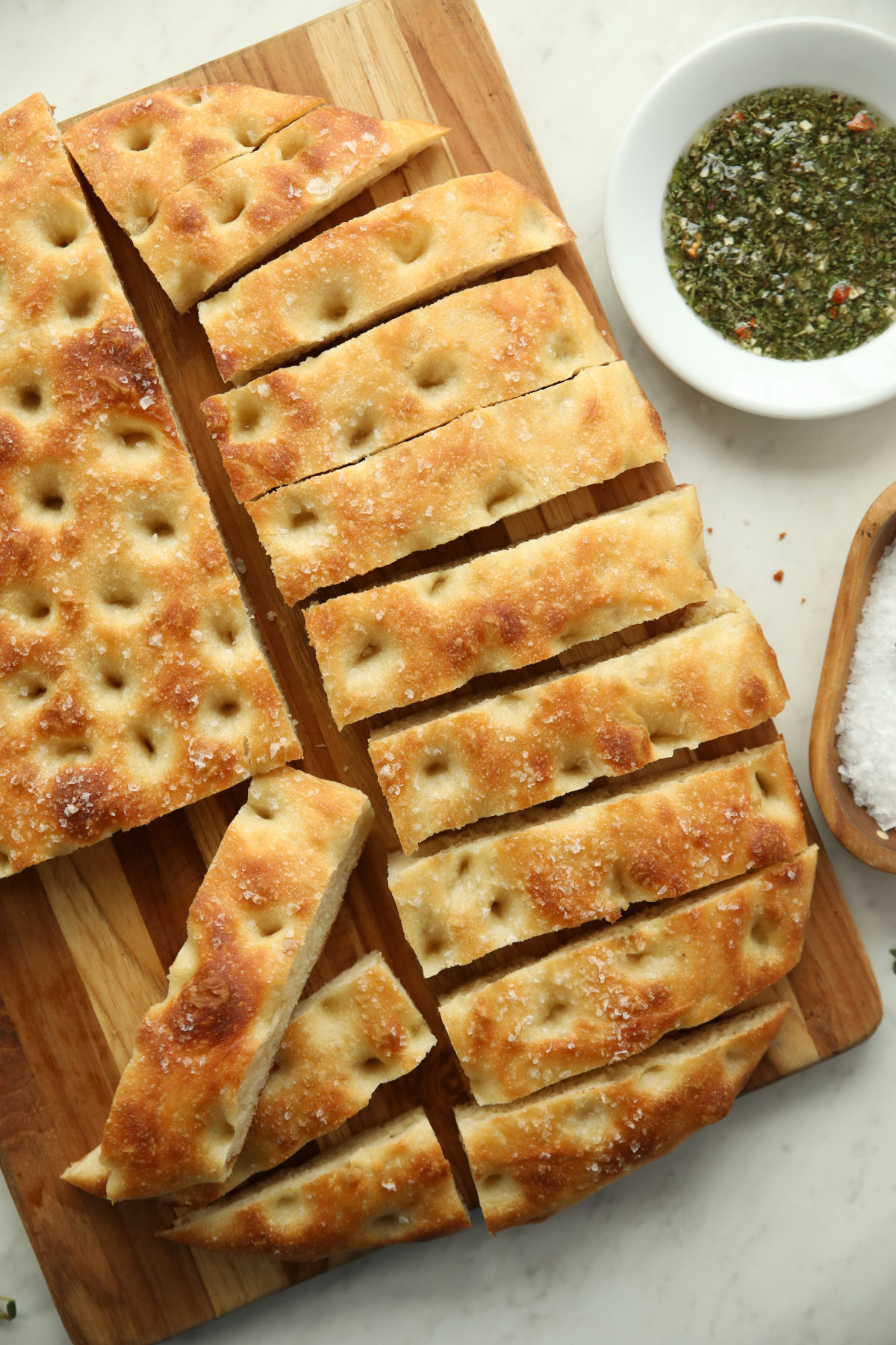 Fresh sourdough focaccia covered in sea salt.  It is in process of being sliced on a cutting board next to dipping oil and sea salt.   