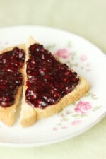 Homemade low sugar blackberry jam spread over whole wheat toast triangles on a white rose china plate sitting on a green table cloth.