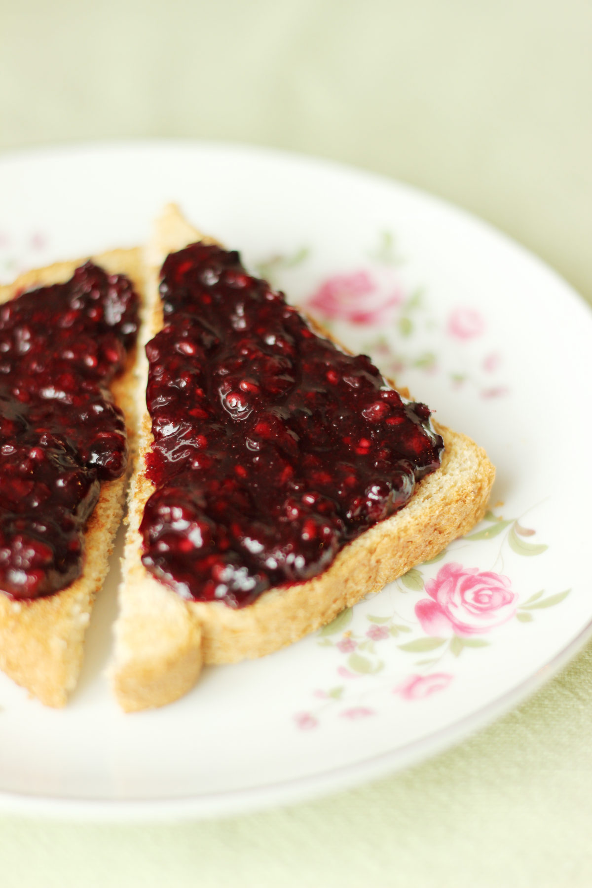Homemade low sugar blackberry jam spread over whole wheat toast triangles on a white rose china plate sitting on a green table cloth. 