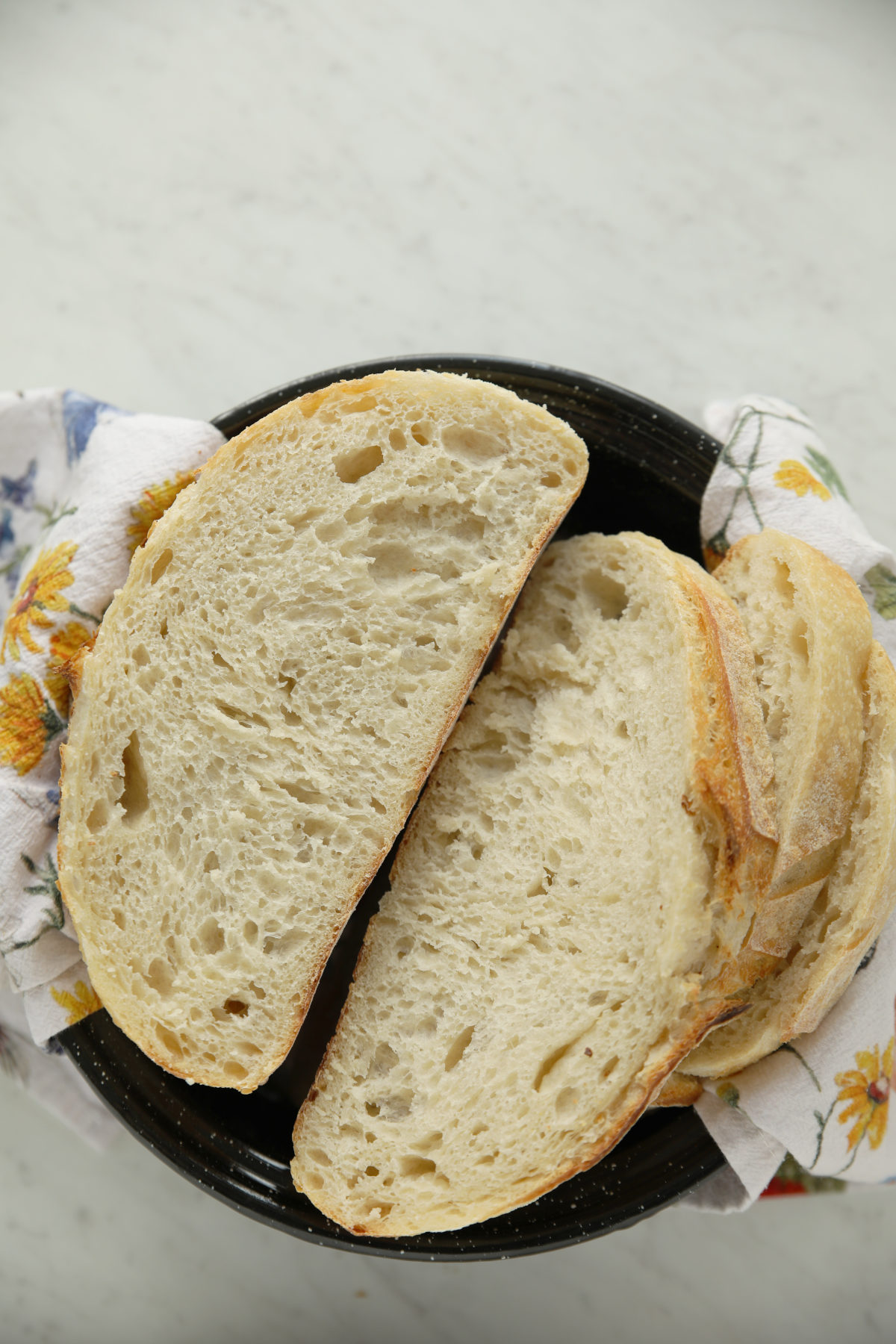 Slices of airy, artisan sourdough bread with a golden crust, arranged in a black speckled graniteware roasting pan lined with a floral tea towel.