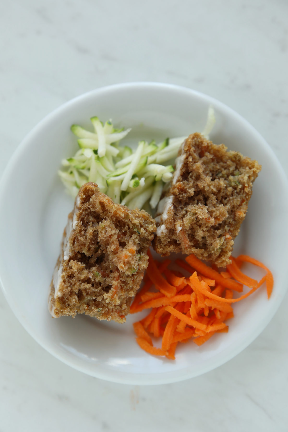 A sourdough discard zucchini carrot muffin split in half on a white plate, showing a tender, moist crumb studded with visible flecks of carrot and zucchini, with shredded carrot and grated zucchini alongside.