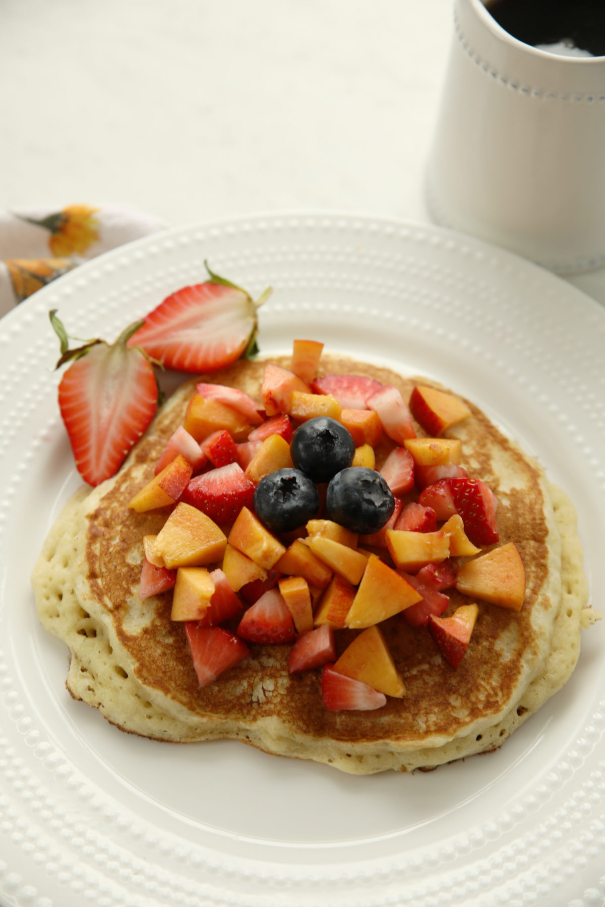 A large sourdough discard pancake topped with diced fresh fruit on a white plate.  You can see strawberries, blueberries and nectarines.   