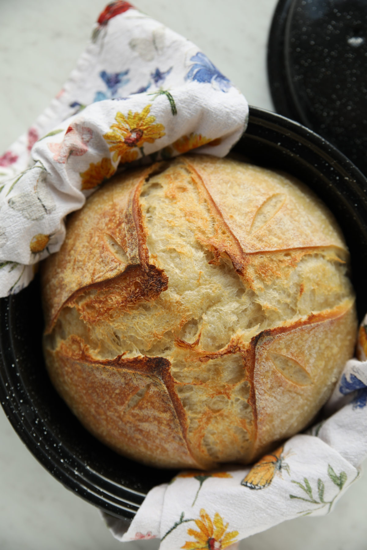 A golden-brown sourdough boule made without a Dutch oven. It has a cross-shaped score and decorative wheat patterns, resting inside a black speckled graniteware roasting pan lined with a floral tea towel.