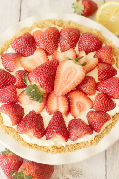A no bake strawberry cheesecake in a white pie plate with a golden graham cracker crust, creamy white cheesecake filling, and fresh sliced strawberries arranged on top, shot from overhead on a white wood surface.