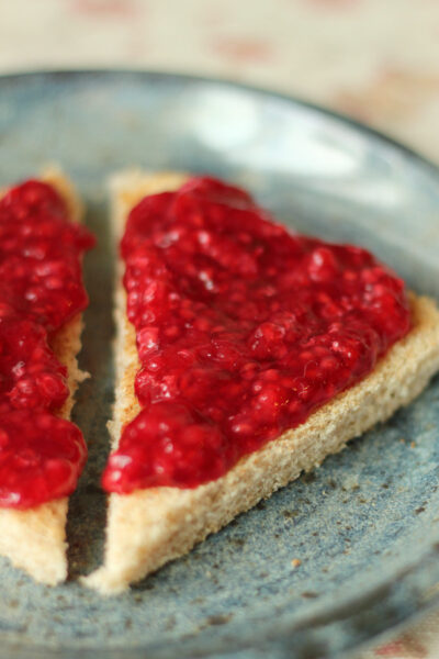 Close up of homemade low sugar raspberry freezer jam spread over whole wheat toast triangles on a blue stoneware plate
