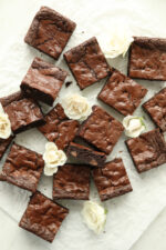 Sixteen fudgy sourdough discard brownies with a crinkly shiny top arranged on white parchment paper with white roses scattered throughout.