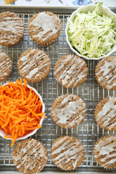 Sourdough discard zucchini carrot muffins with apple cider glaze drizzled on top, cooling on a wire rack with bowls of shredded zucchini and fresh grated carrot alongside.