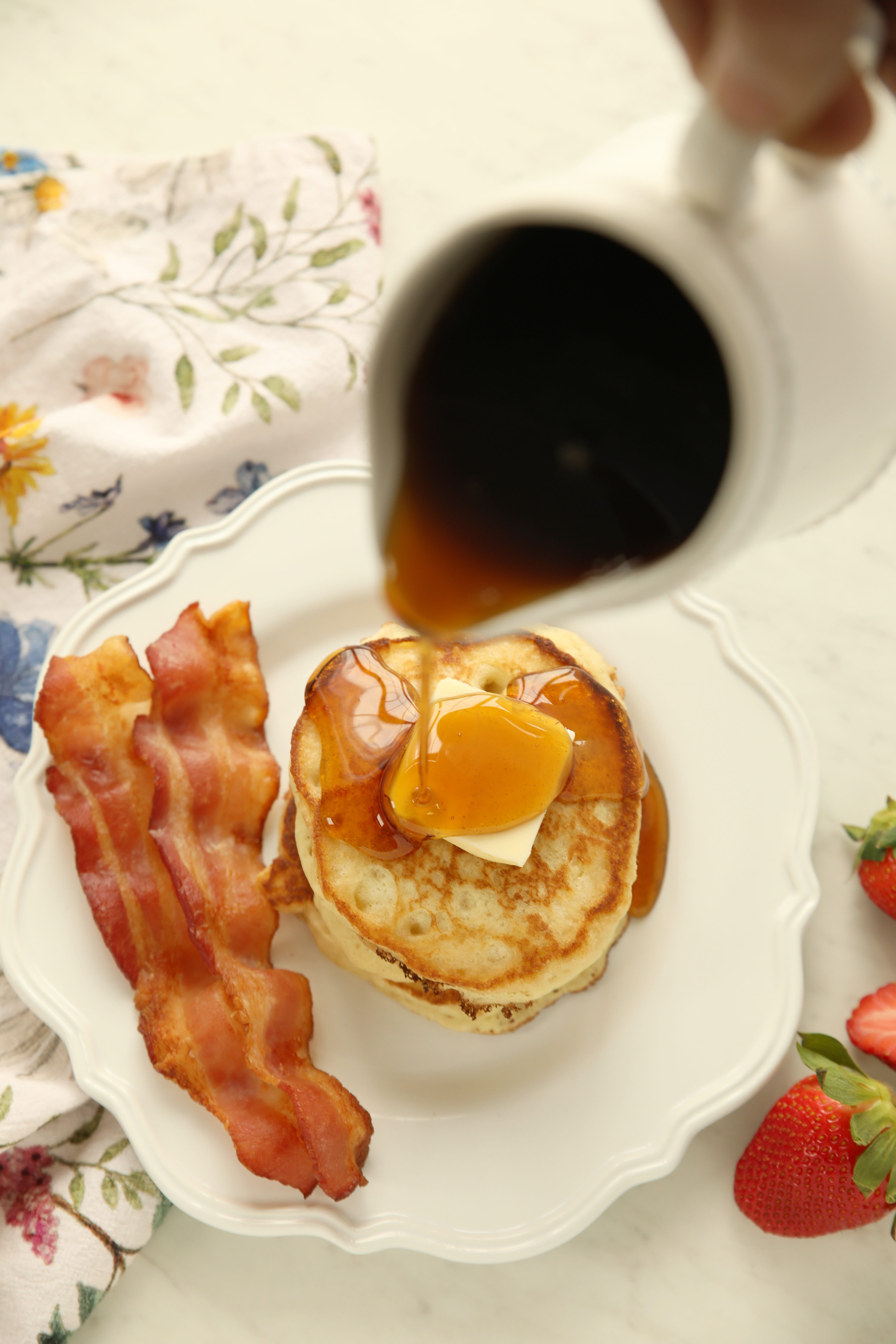 A pitcher of maple syrup pours on top of a stack of sourdough discard pancakes.  You can see bacon on the plate and butter on the pancakes. There are strawberries and a floral tea towel nearby.