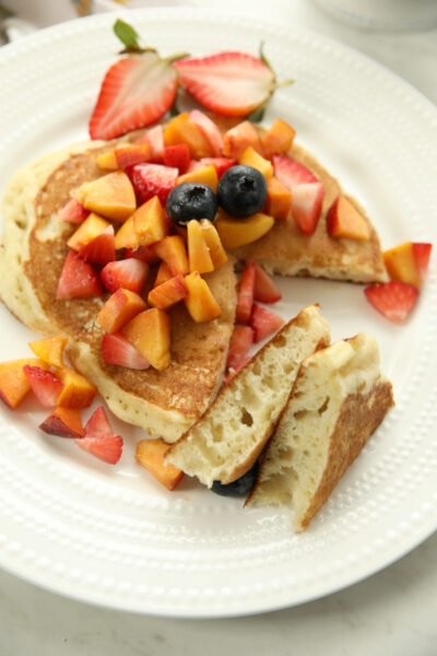 A fluffy sourdough discard pancake on a white beaded plate, topped with fresh diced peaches, strawberries, and blueberries. A cut slice is propped up to show the light, airy interior crumb of the sourdough discard pancake recipe.