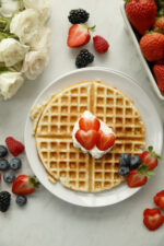 A stack of golden sourdough discard waffles on a white plate topped with sliced fresh strawberries, surrounded by whole strawberries and blueberries on a marble surface.