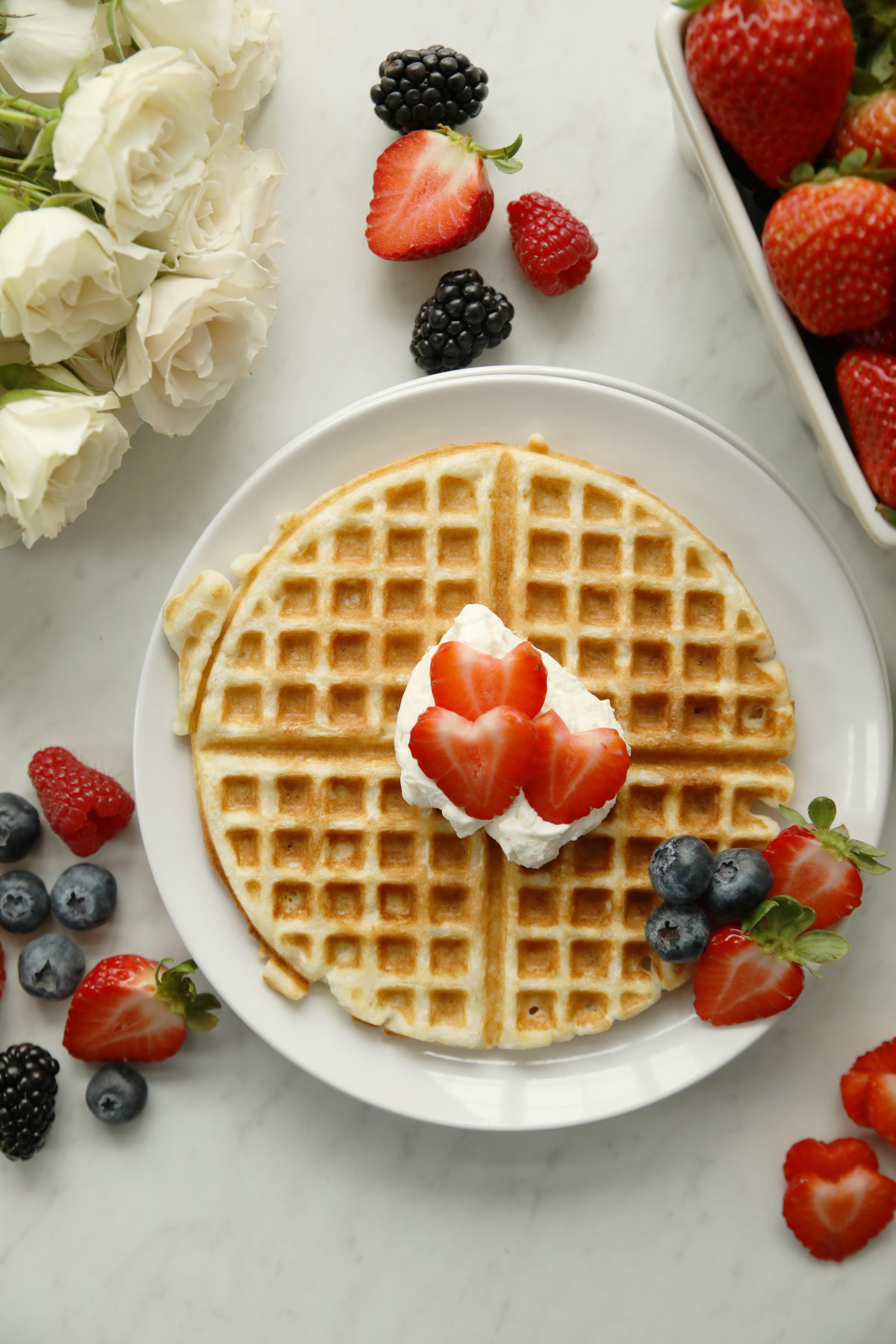 A golden sourdough discard waffle on a white plate topped with whipped cream and sliced strawberries, surrounded by fresh strawberries, blueberries, and blackberries on a marble surface with white roses nearby.