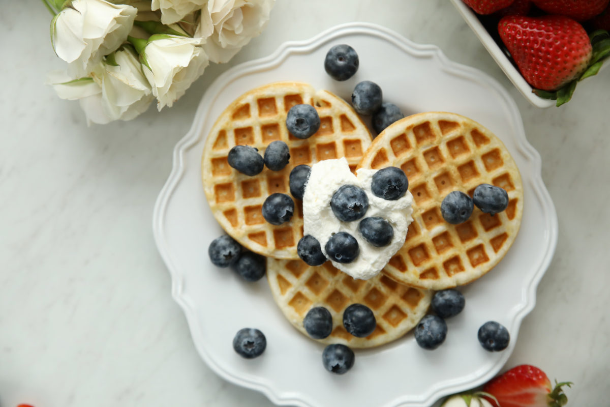 Two golden sourdough discard waffles on a white scalloped plate topped with whipped cream and fresh blueberries, with white roses and strawberries nearby on a marble surface.