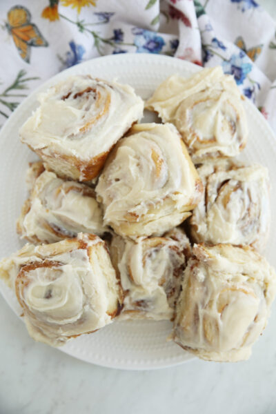A white plate piled with frosted sourdough cinnamon rolls showing golden swirls and thick buttercream glaze, with a floral napkin in the background.