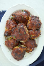 Oven BBQ chicken thighs piled high on a white oval platter, coated in deep caramelized BBQ sauce and scattered with fresh chopped parsley.