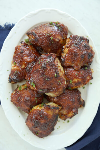 Oven BBQ chicken thighs piled high on a white oval platter, coated in deep caramelized BBQ sauce and scattered with fresh chopped parsley.