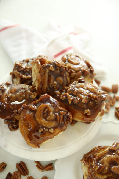 Sourdough discard sticky buns with pecans on a white cake stand, drizzled with caramel brown sugar syrup