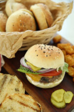 Sourdough hamburger buns with buttermilk on a wooden board, one bun loaded with a cheeseburger, lettuce, tomato, pickles, and red onion, with a basket of buns lined with brown parchment in the background.