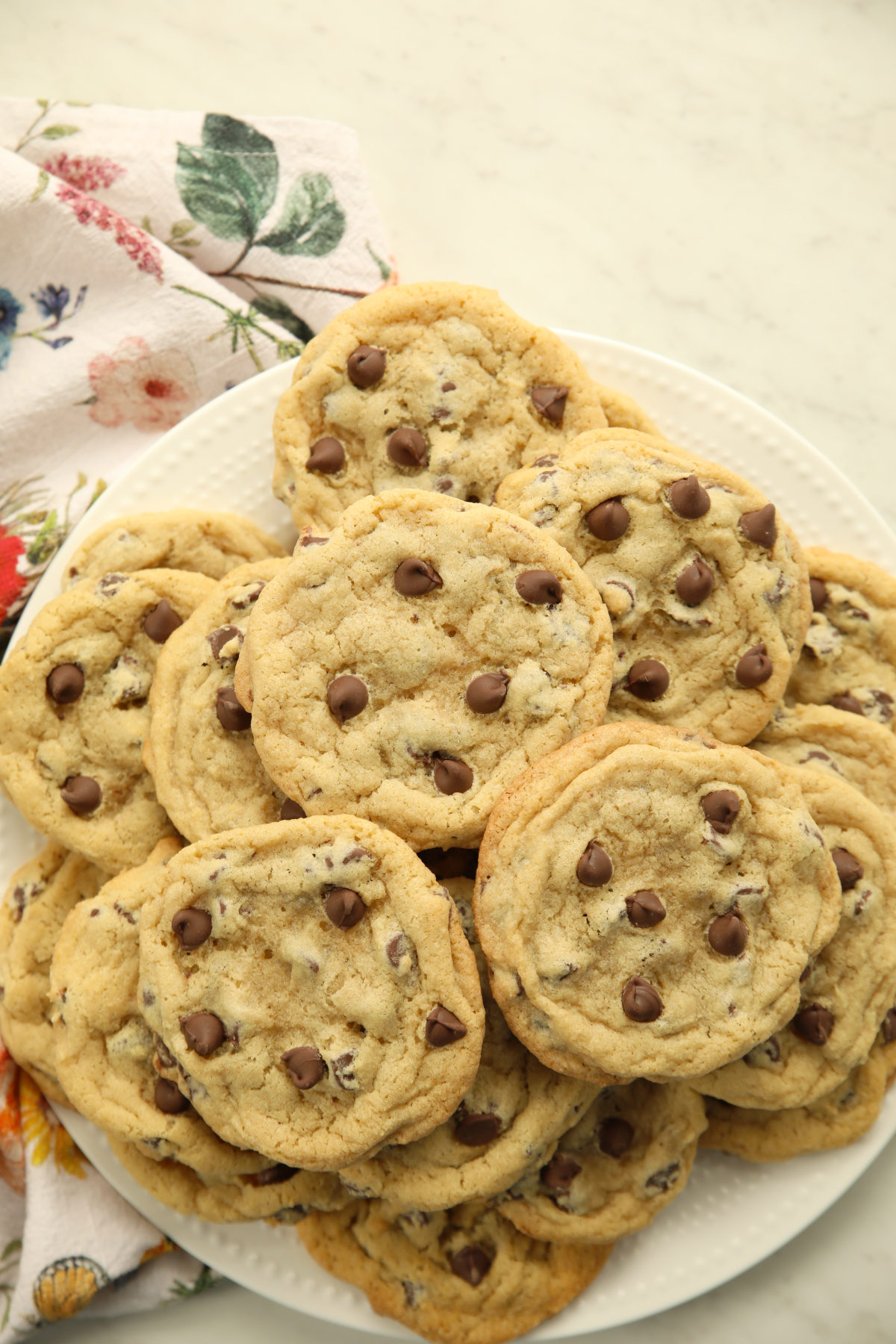 A plate piled high with chewy sourdough discard chocolate chip cookies on a white marble surface with a floral tea towel.